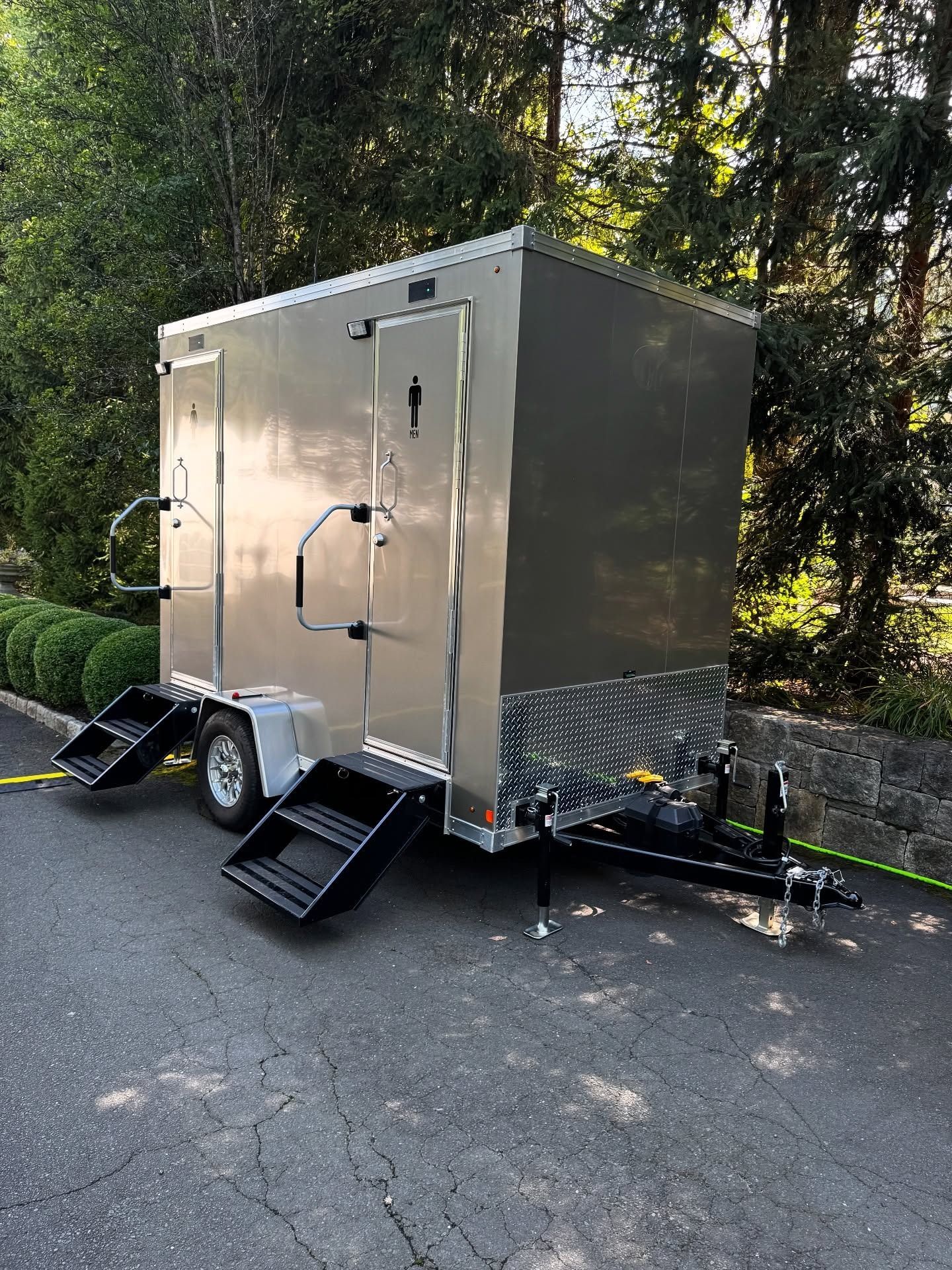 Silver mobile restroom trailer on asphalt, surrounded by greenery. Black steps, two doors.