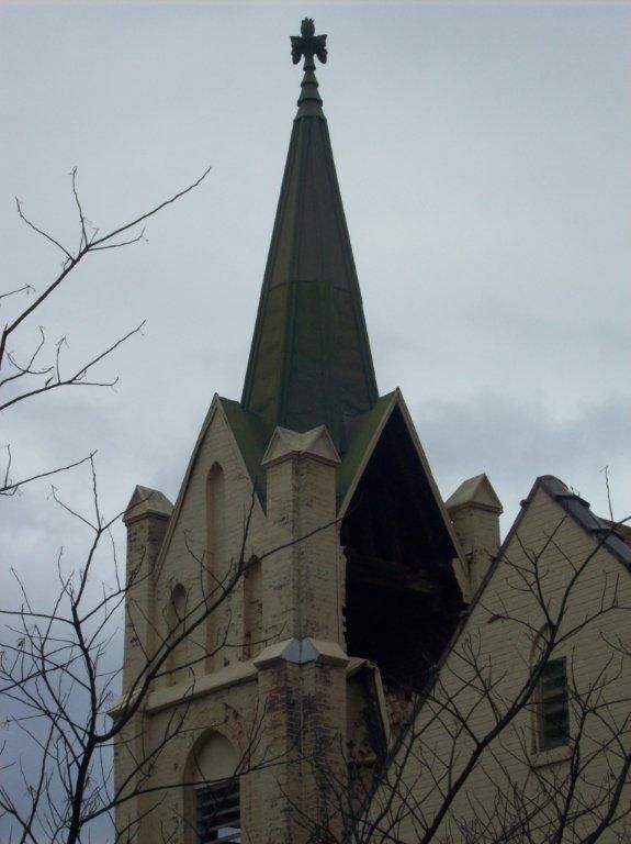 Church steeple with a cross against a cloudy sky, viewed above bare tree branches