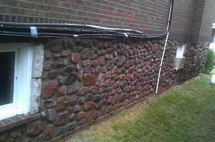 Basement wall with irregular red and gray stone facing beside a white-framed window and grassy yard