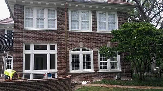 Brick two-story house with white-trim windows, a front lawn, and a person in yellow bending near the left side.