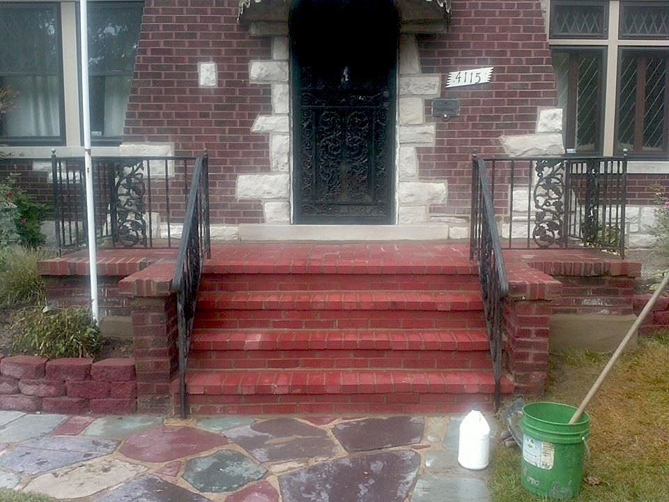 Front steps of a brick house with red stairs, black railings, and a green bucket on the right
