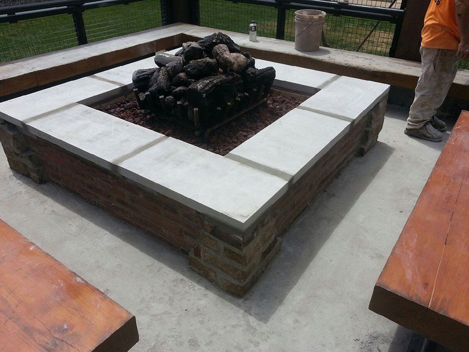 Stone fire pit with white benches in an outdoor seating area; a person stands nearby.