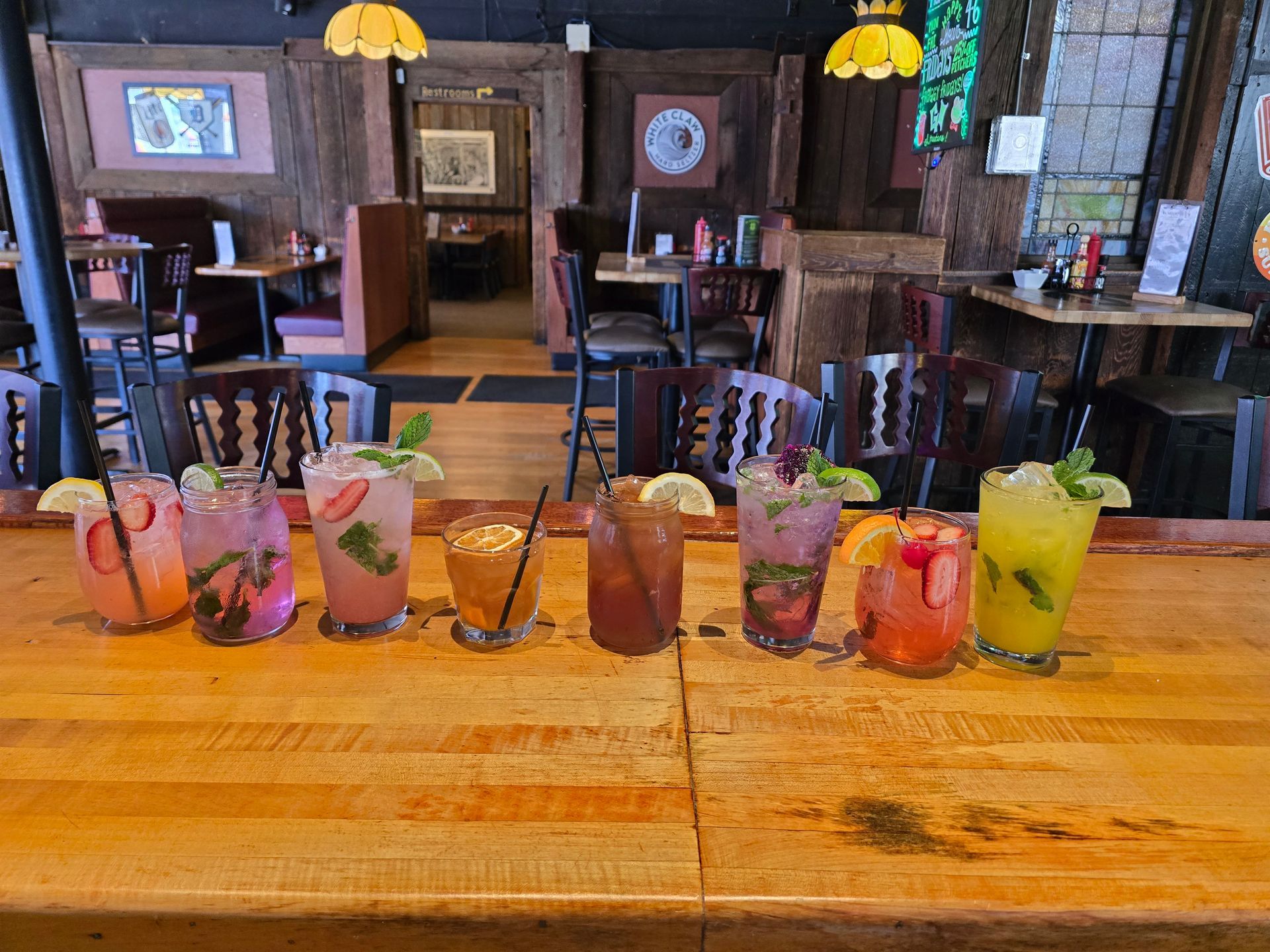 A row of eight colorful cocktails in varied glasses sits on a wooden bar in a dimly lit restaurant.