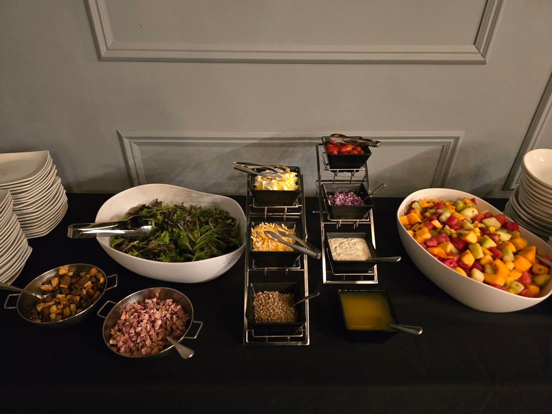 A salad bar with bowls of greens, fruit, and various toppings on a black tablecloth against a light wall.