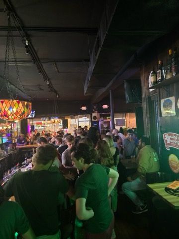 A crowded, dimly lit bar with patrons standing around, featuring a stained-glass lamp and wall decorations.