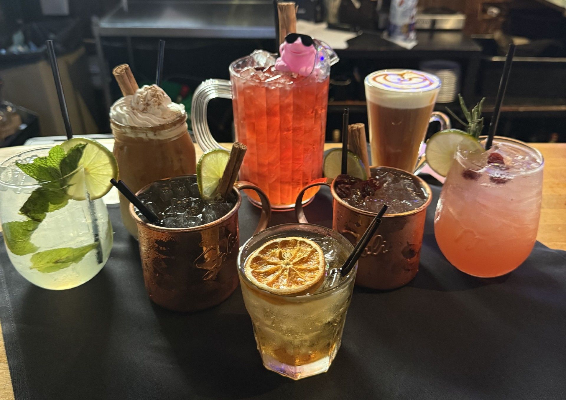 Five colorful cocktails with garnishes lined up on a wooden bar in a dimly lit restaurant.