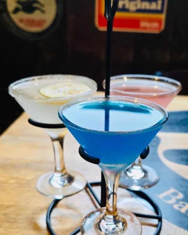 Three martini glasses with blue, pale yellow, and pink cocktails in a metal stand on a wooden table.