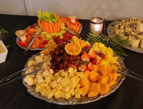 A platter of cubed cheese, grapes, and strawberries sits next to a veggie tray and pinwheels on a dark tablecloth.