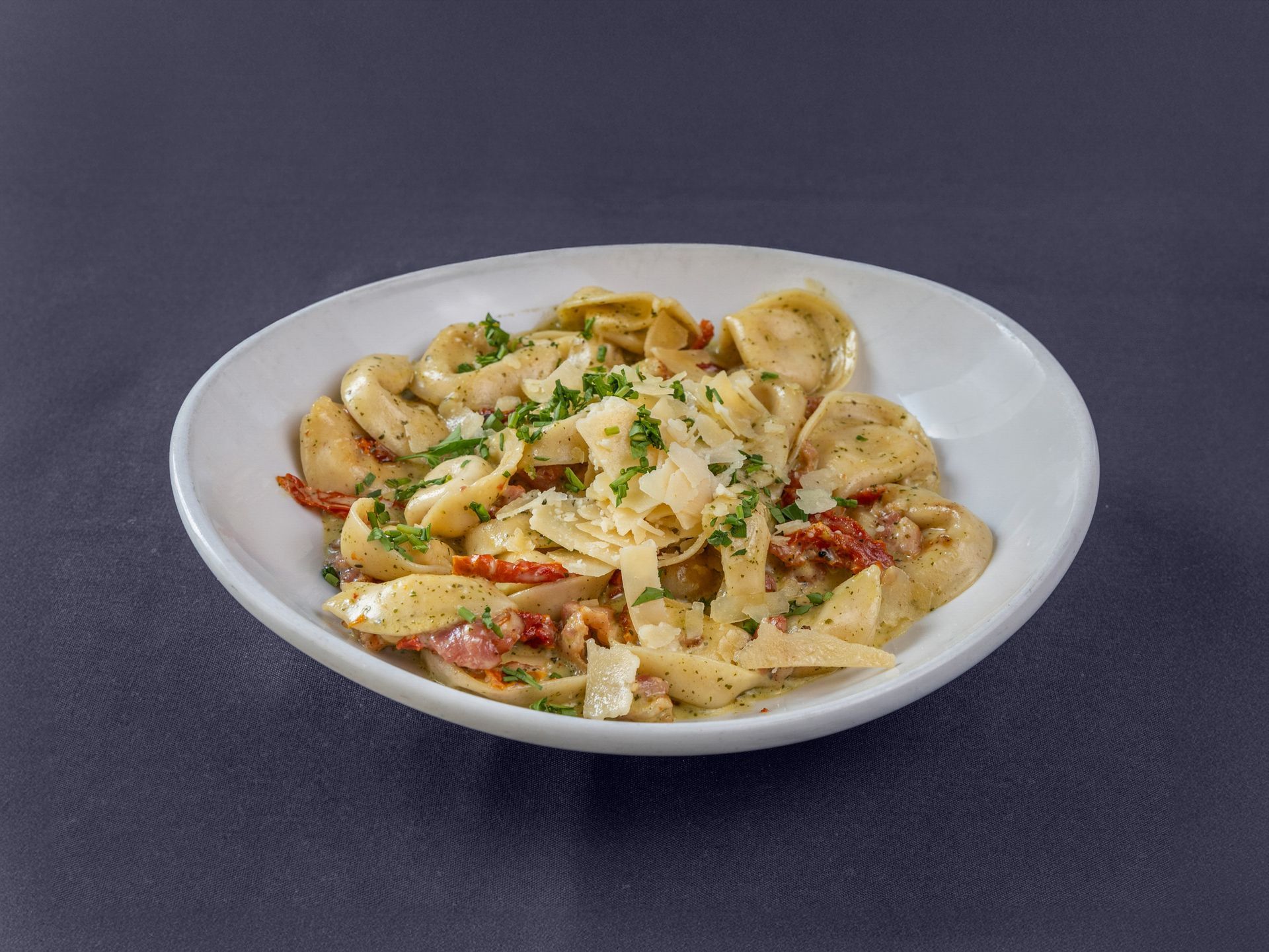 A white bowl filled with creamy pasta, garnished with sun-dried tomatoes, herbs, and shaved parmesan cheese.