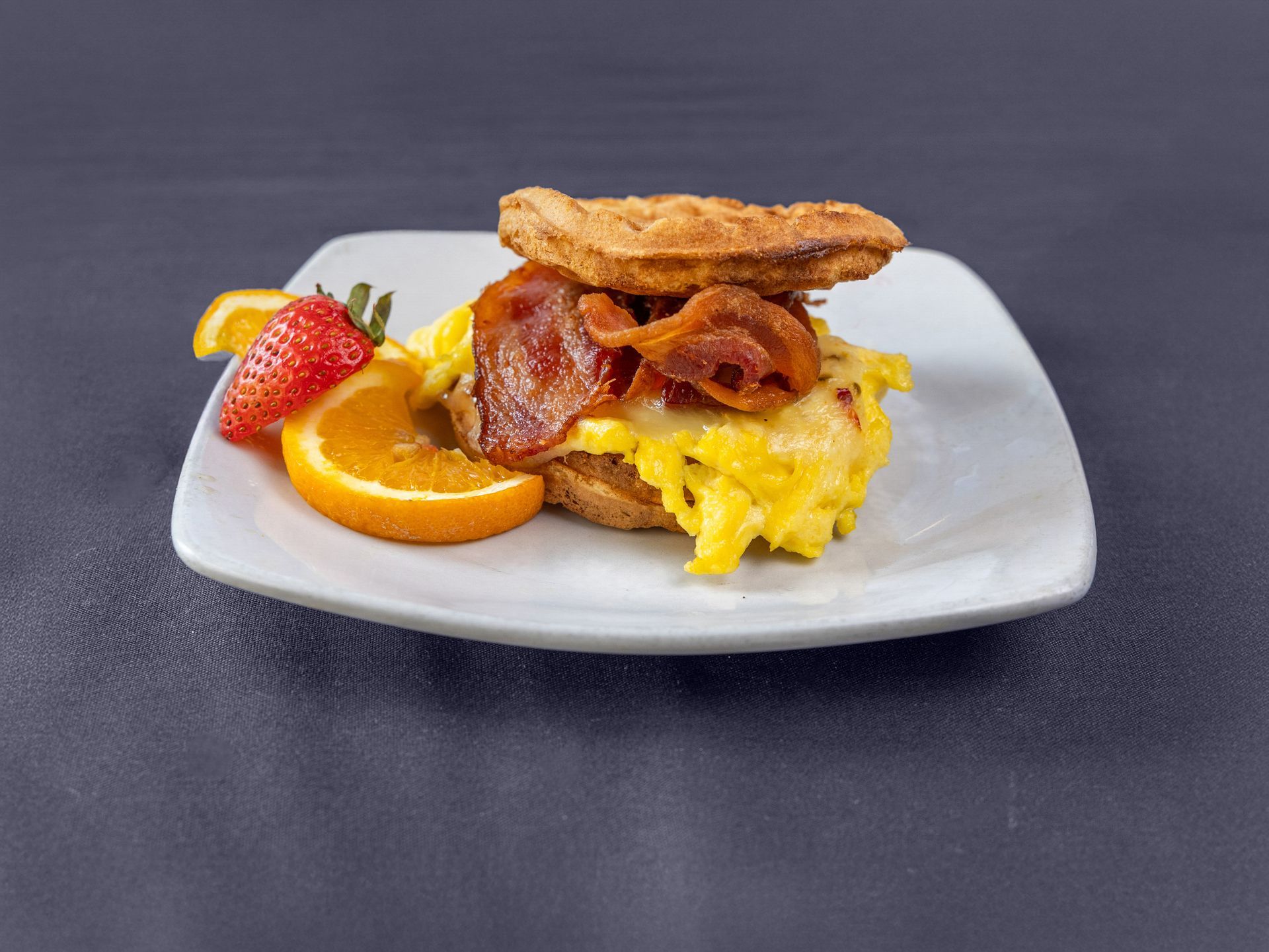 A breakfast sandwich with scrambled eggs and bacon on a waffle, served with a strawberry and orange slice on a white plate.