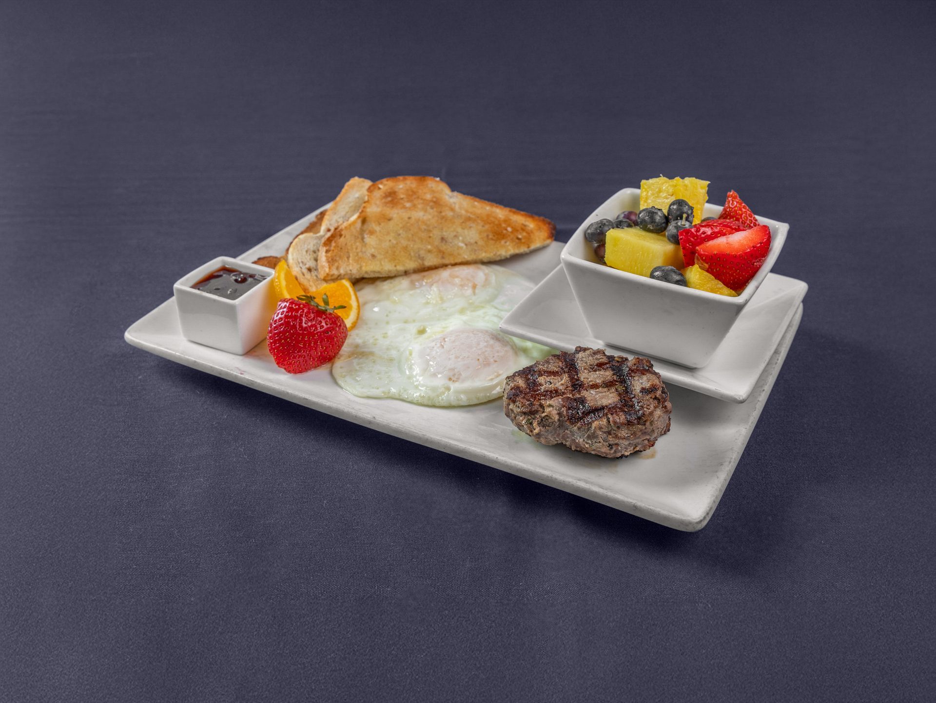 A white rectangular plate features two fried eggs, a grilled sausage patty, toast, a strawberry, and a bowl of fruit salad.