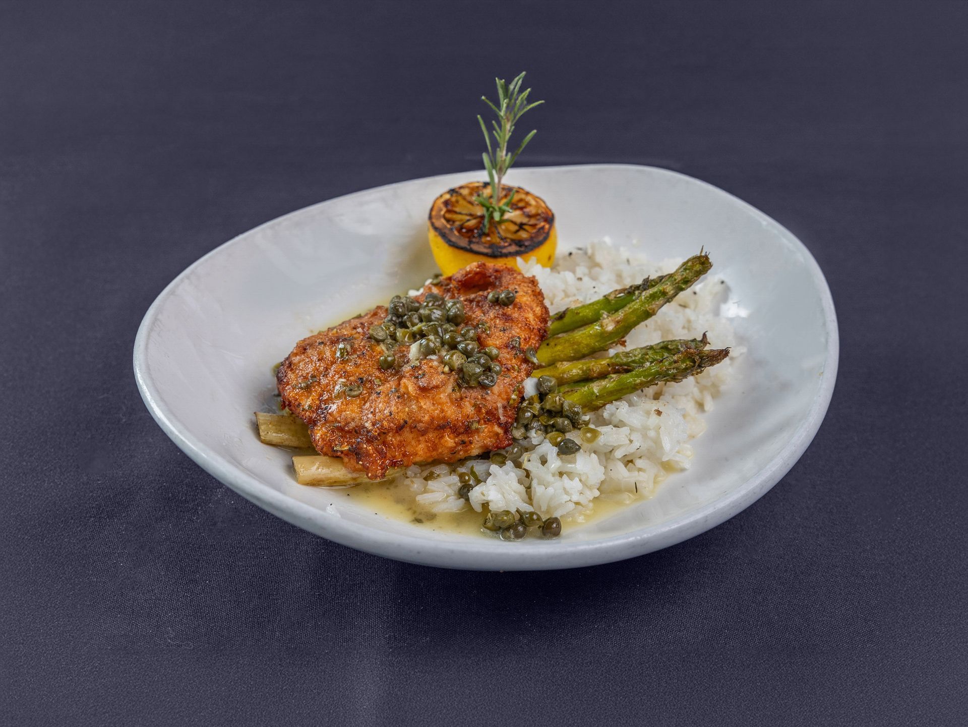 A plated dish of golden-brown chicken with caper sauce, served alongside white rice, asparagus, and a charred lemon slice.