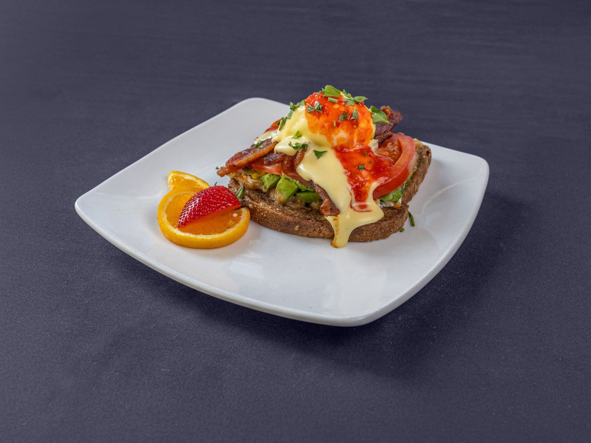 An open-faced sandwich topped with a poached egg, hollandaise, bacon, and tomato on a white plate with orange and berry.