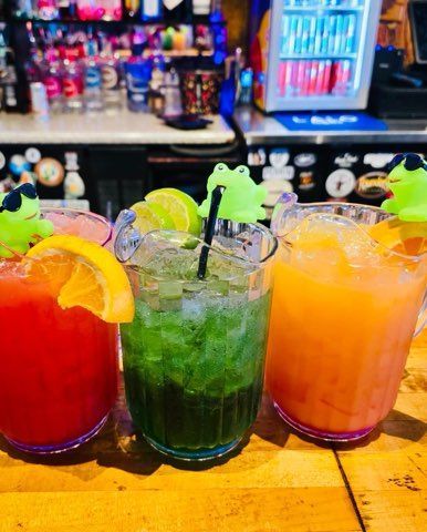 Three cocktails with lime and orange garnishes and decorative frog toppers sit on a wooden bar in a dimly lit venue.