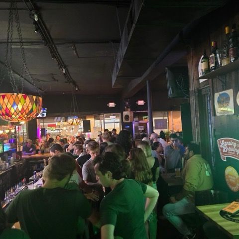 A crowded, dimly lit bar with patrons gathered at the counter and tables, featuring stained-glass lamps overhead.