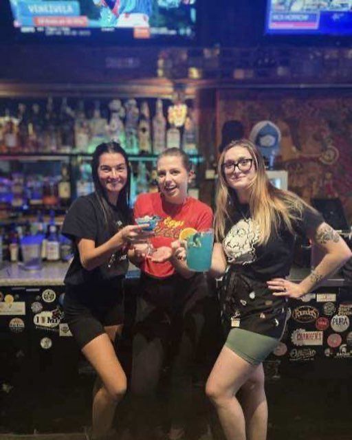 Three people stand behind a bar, smiling and holding blue drinks, with a background of shelves filled with bottles.