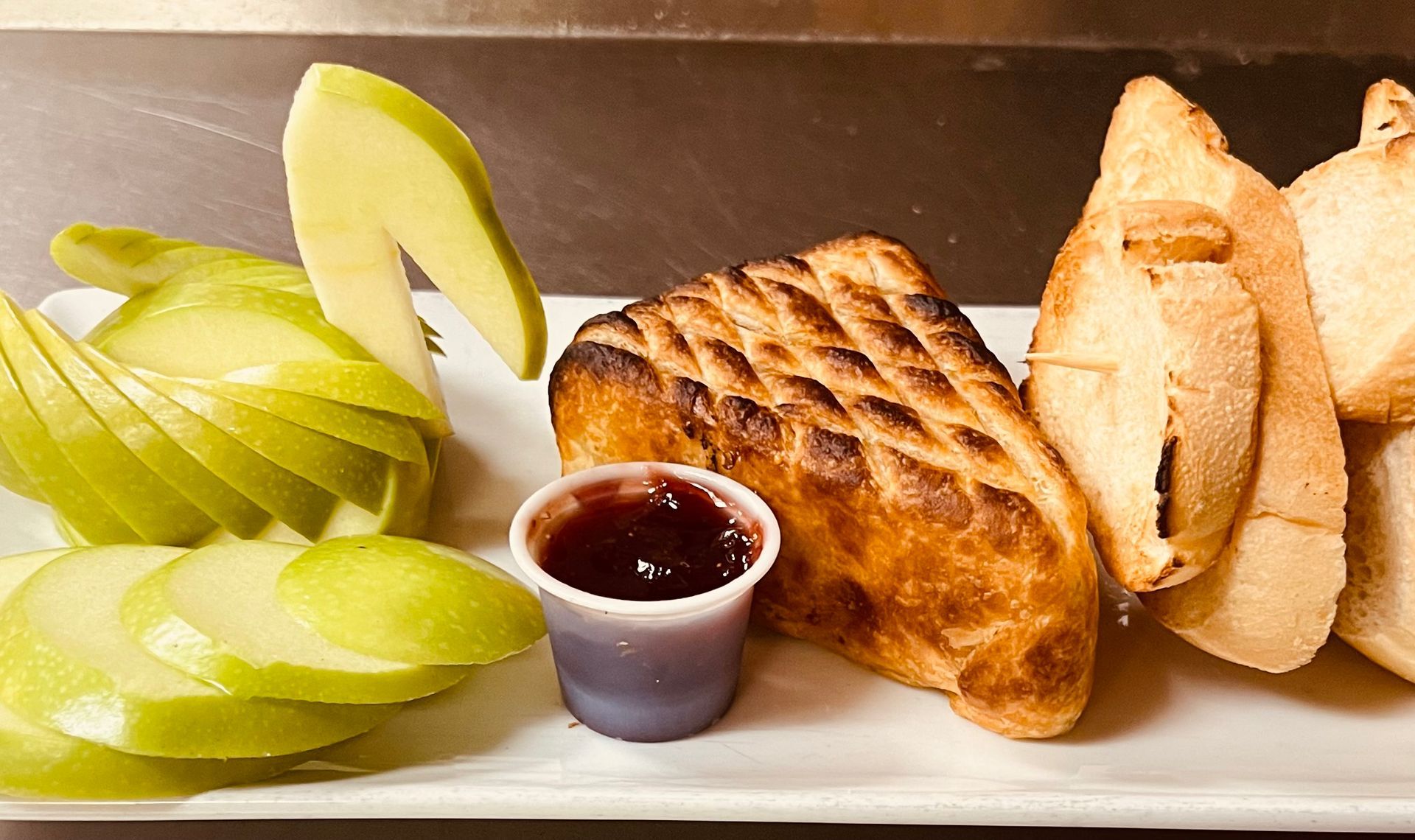 A plate featuring a grilled sandwich, sliced green apples, a small cup of berry jam, and toasted baguette slices.