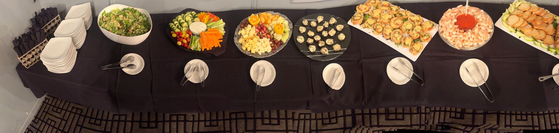 A buffet table with a black tablecloth features a salad, vegetable tray, cheese platter, appetizers, and snacks.