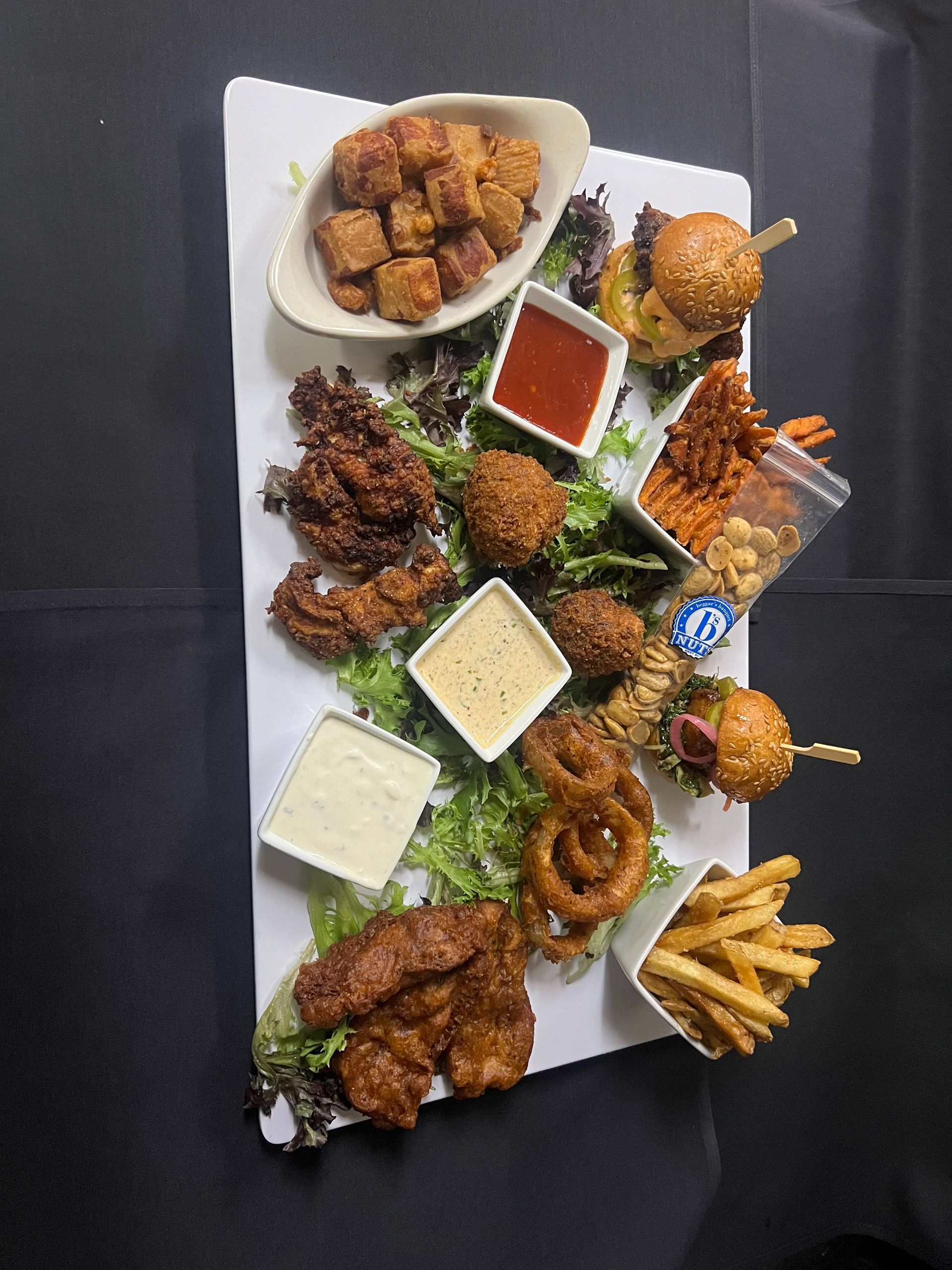 A large rectangular platter of assorted fried appetizers, including sliders, onion rings, fries, and dipping sauces.