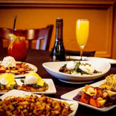 A table spread featuring various brunch dishes, including eggs Benedict, roasted vegetables, a bottle, and cocktails.