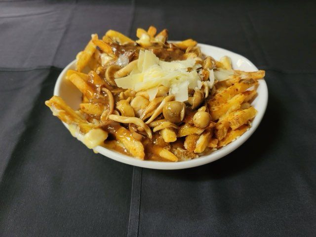 A bowl of poutine topped with sautéed mushrooms and shaved cheese on a black surface.