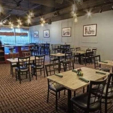 A restaurant dining room with square wooden tables, black chairs, patterned carpet, and hanging lights.