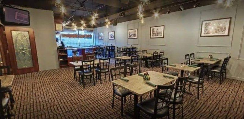 A dining area with wooden tables, black chairs, and patterned carpet under warm overhead lighting.