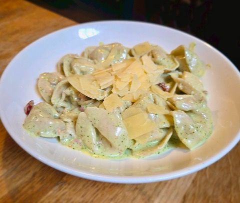 A white bowl filled with tortellini in a creamy green sauce, topped with shaved parmesan cheese on a wooden table.