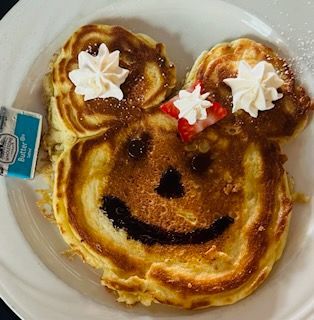 A Mickey Mouse-shaped pancake on a white plate with whipped cream dollops, fruit, a chocolate face, and a butter packet.