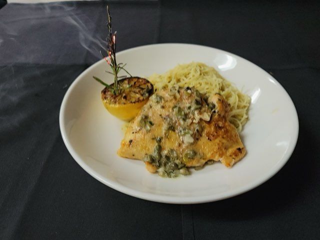 A plated dish of chicken topped with a caper sauce, served alongside pasta and a roasted lemon half on a black surface.