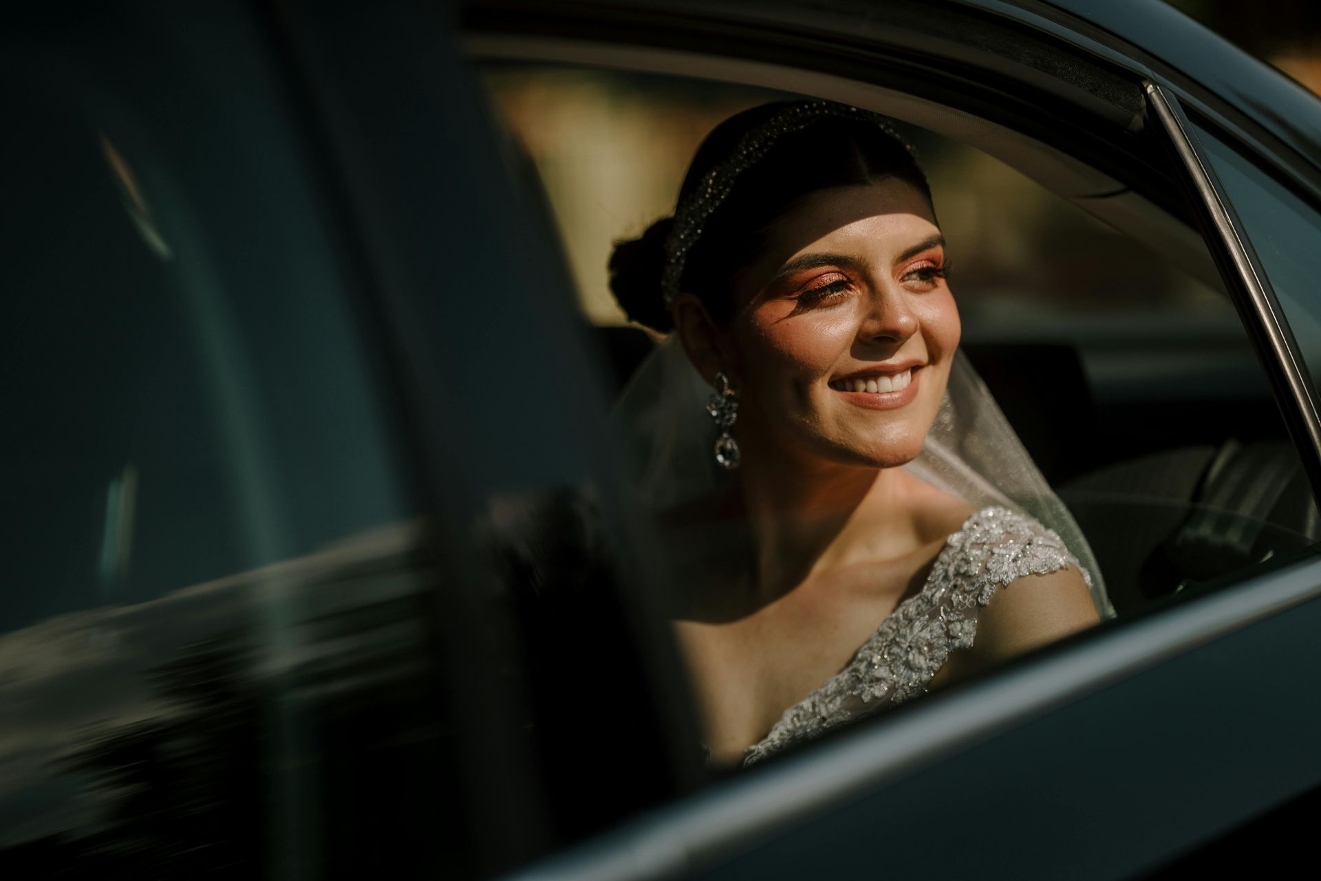 Bride smiling in a car, wearing a white dress and veil, with sunlight on her face.