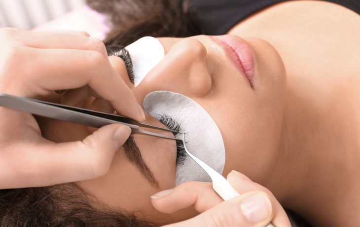 Close-up of eyelash extensions being applied with tweezers and white under-eye pads