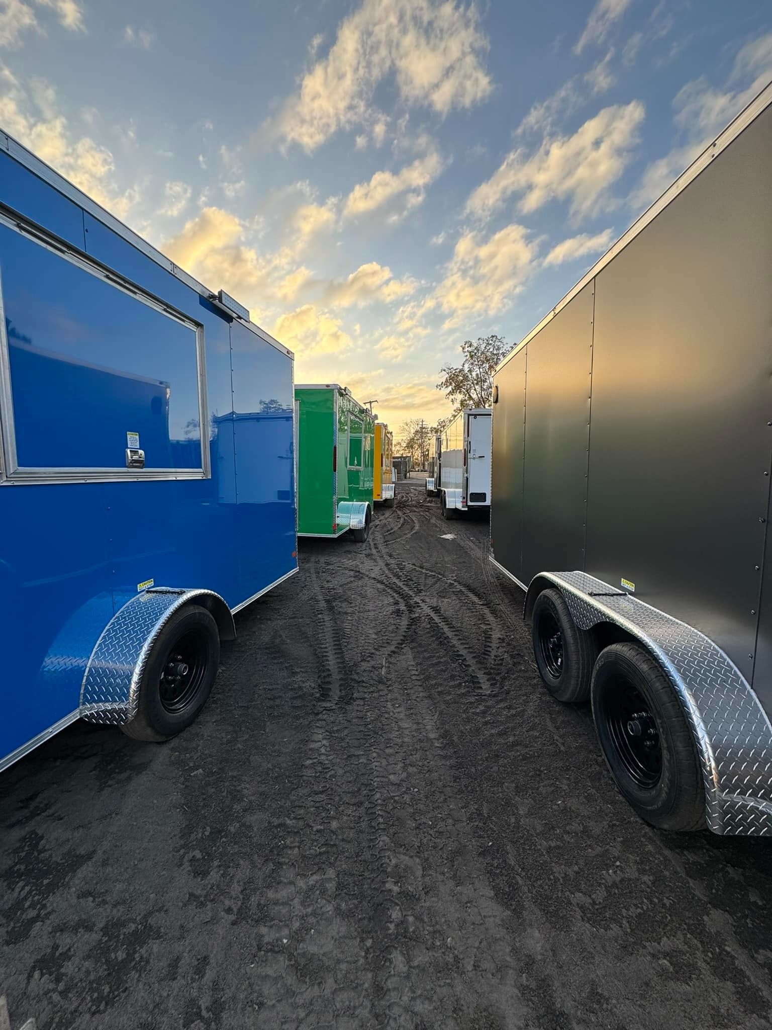 Food trailers parked on dirt, various colors, under a cloudy sky.