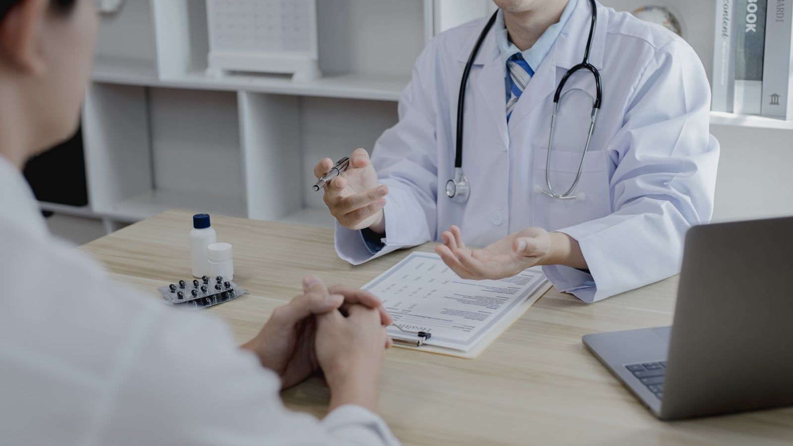 Doctor consulting a patient at a desk, holding a pill bottle beside a laptop.