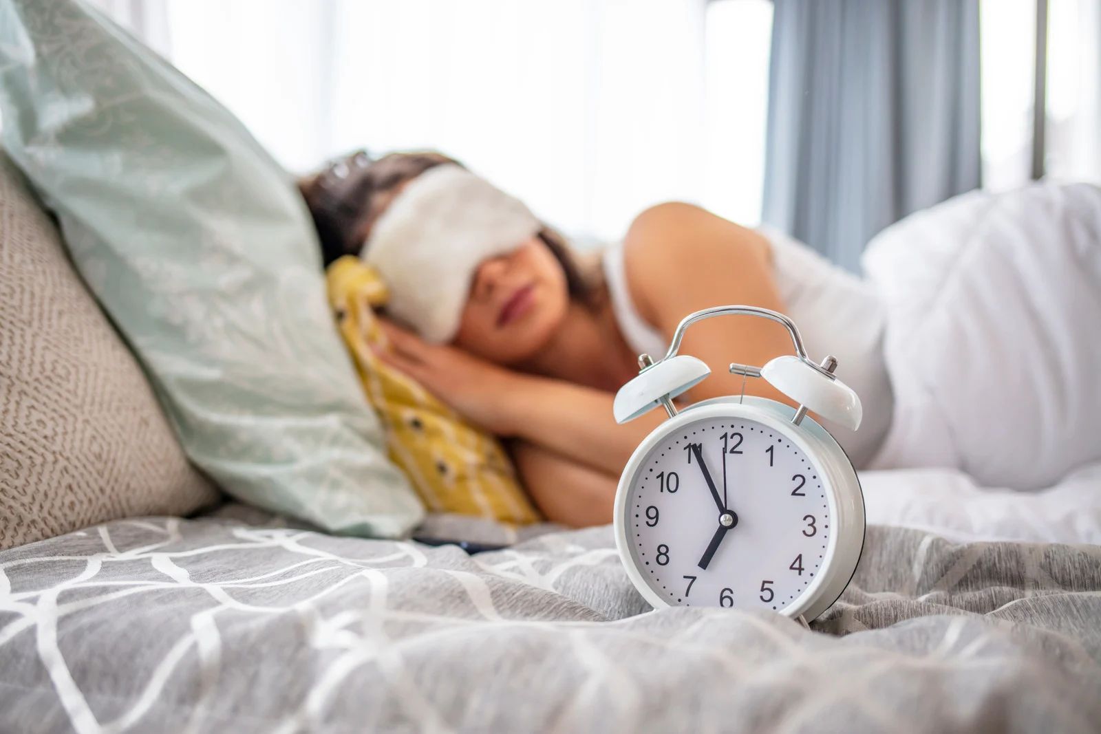 A person wearing a sleep mask rests in bed near a white alarm clock that shows the time is shortly after seven.