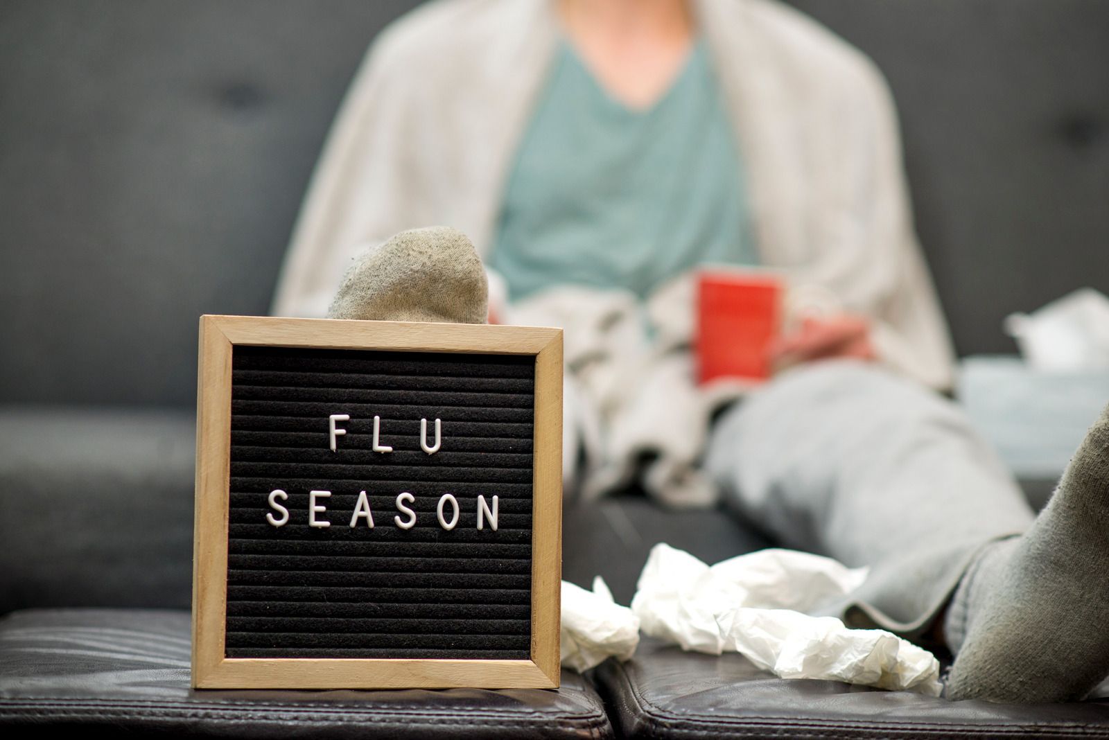Sign reading “FLU SEASON” on a couch beside tissues, a blanket, and a red mug