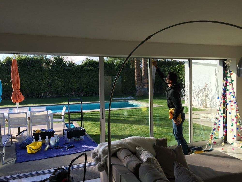 Person hanging decorations on a patio beside a pool, seen through open glass doors.
