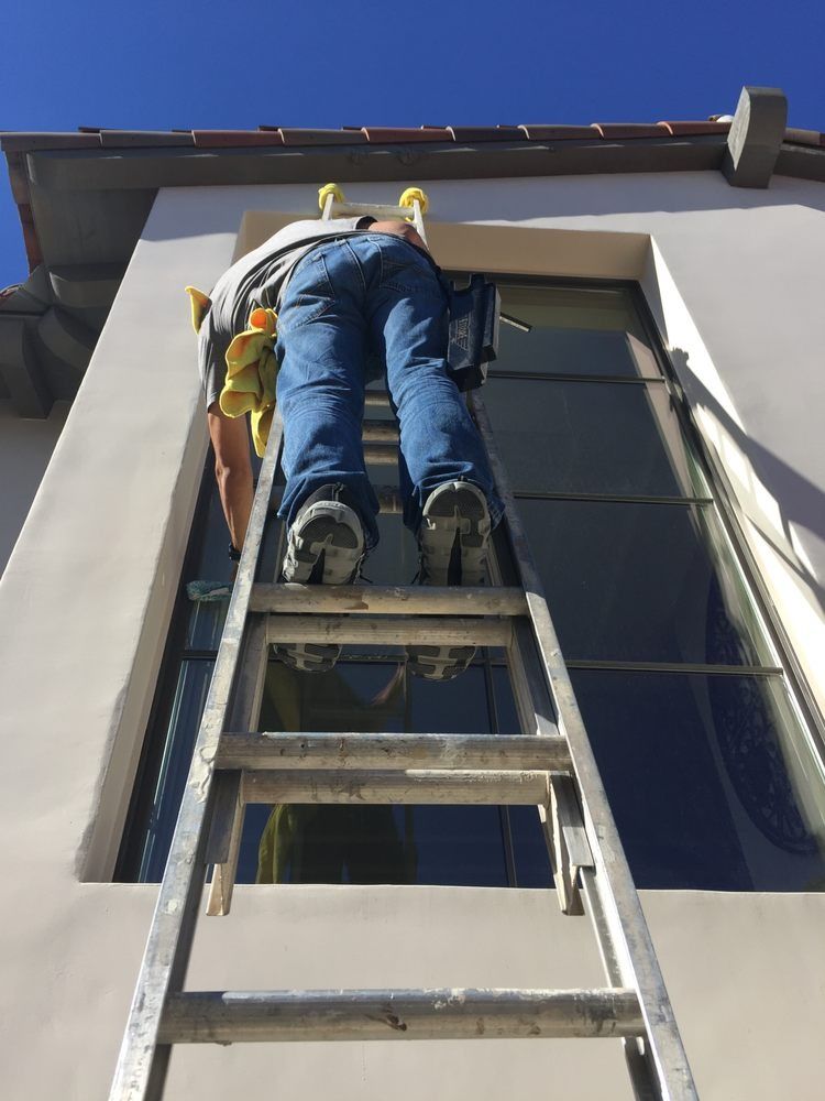 Worker on a ladder painting or repairing the exterior of a building beneath a window.