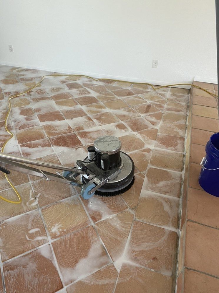 Floor buffer polishing a tiled floor beside a white wall and a blue bucket.