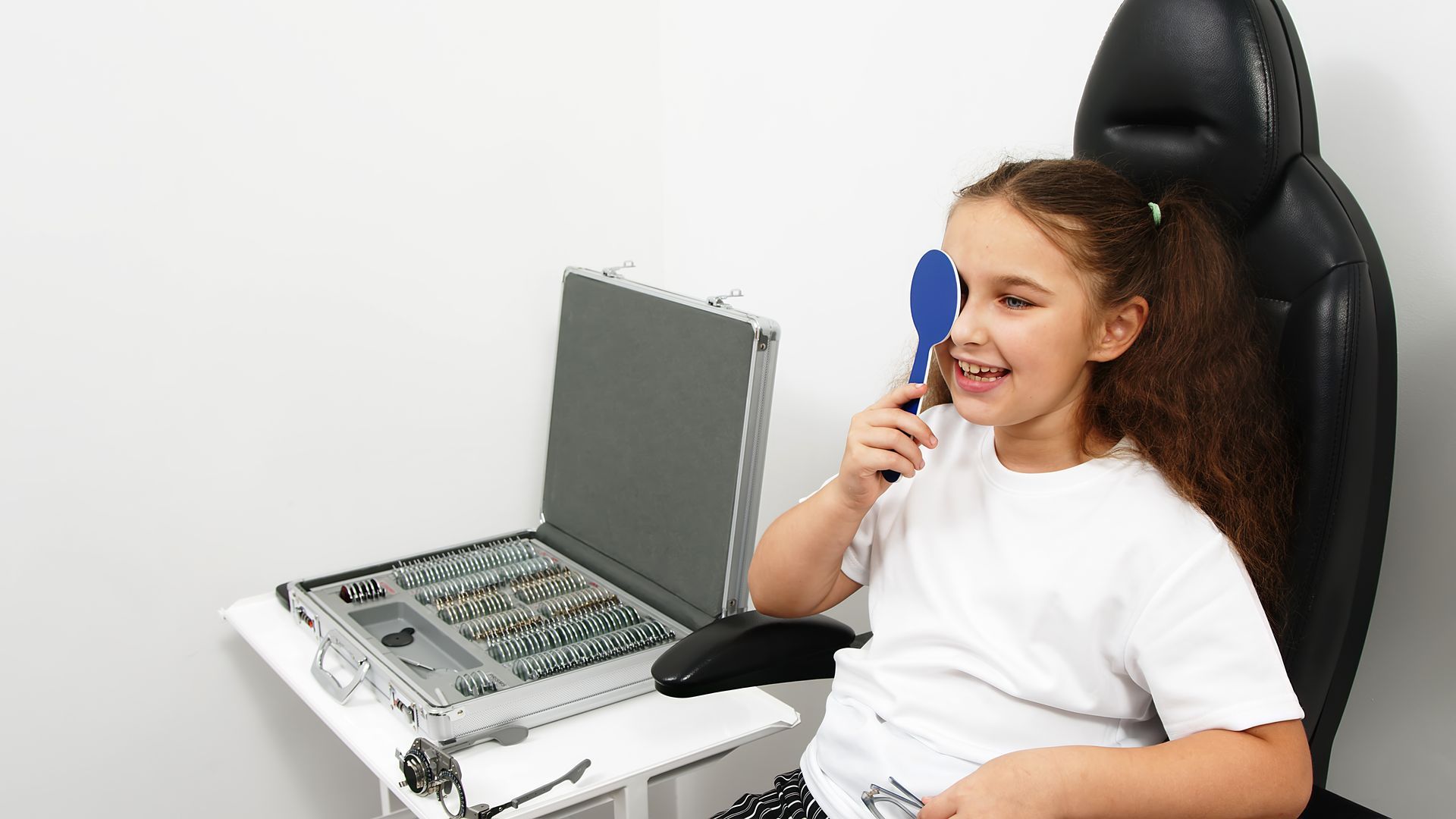 Smiling girl covering eye with occluder paddle during vision test at optometrist's office.