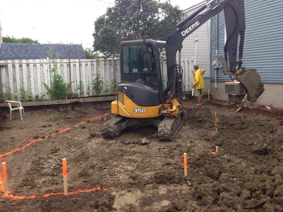 Une excavatrice Deere creuse dans la terre devant une maison