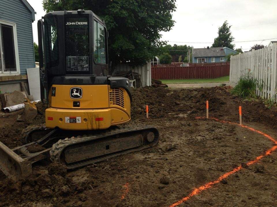 Une excavatrice jaune John Deere creuse dans la terre devant une maison.