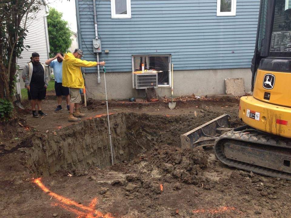 Un homme en veste jaune mesure un trou dans le sol devant une maison.