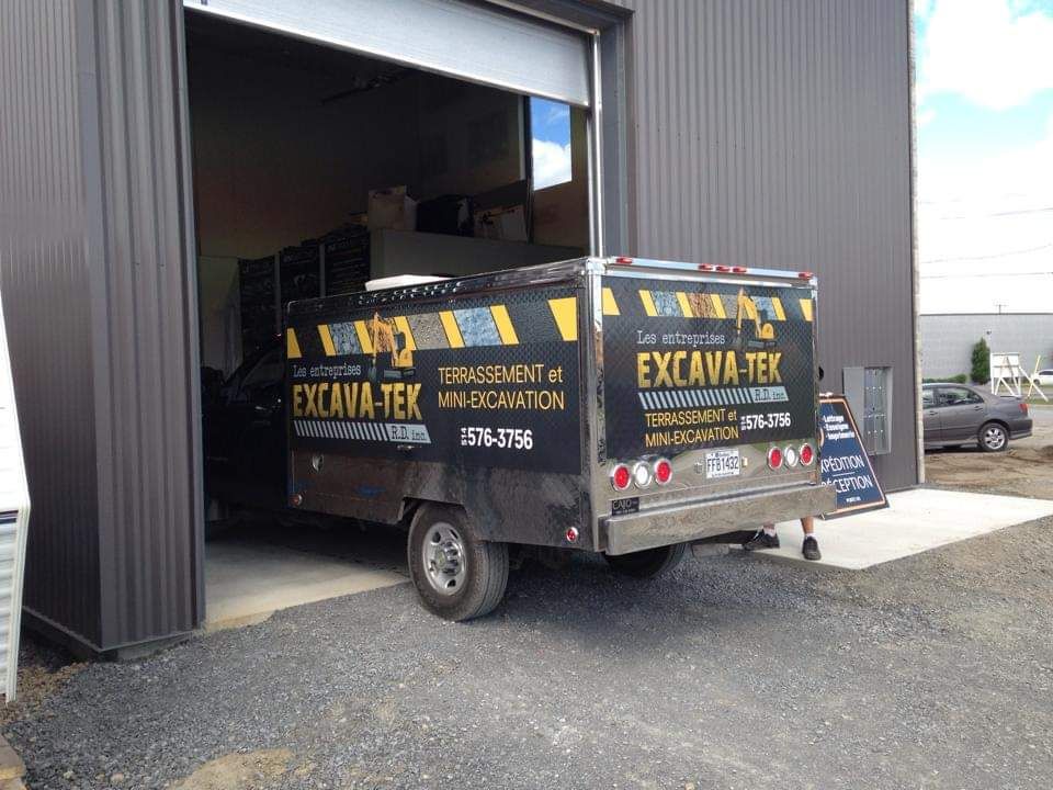 Un camion noir avec excava tek écrit à l'arrière est garé devant un bâtiment.