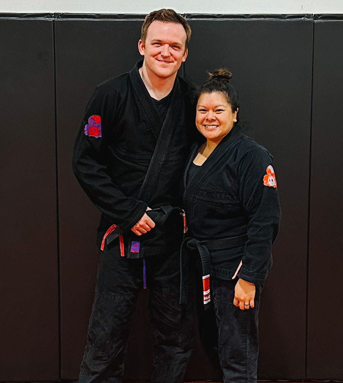 Two smiling martial artists in black gis stand side-by-side against a black wall mat.