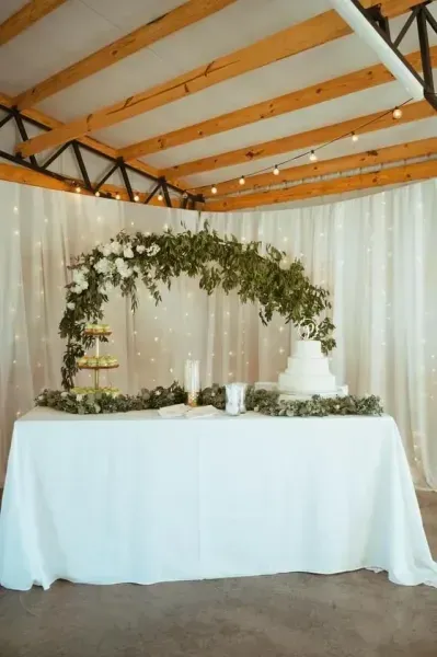 Decorated dessert table with white draping and greenery arch in an indoor event venue