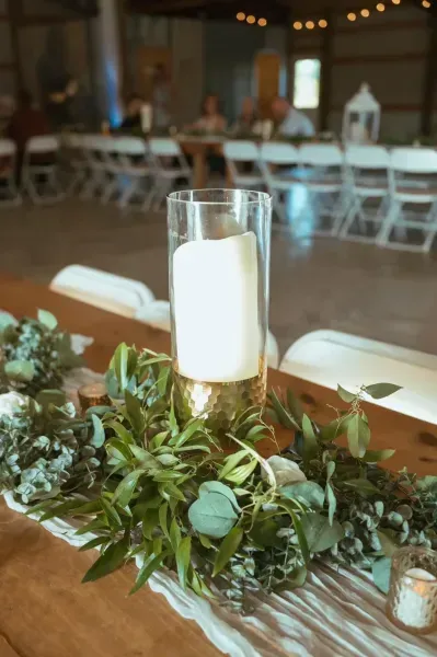 Table centerpiece with greenery and a tall white candle in a gold holder at a softly lit event venue