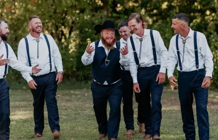 Five men in suits and suspenders laughing outdoors at a garden event