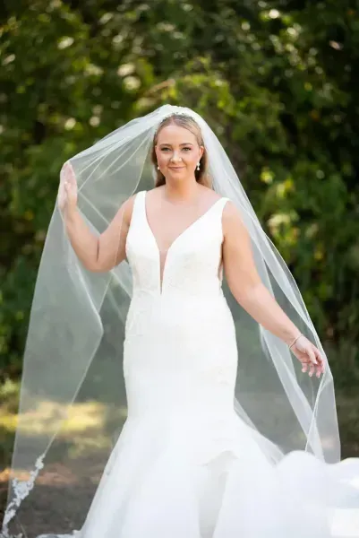 Bride in a white wedding gown and veil standing outdoors, holding the veil against a leafy background