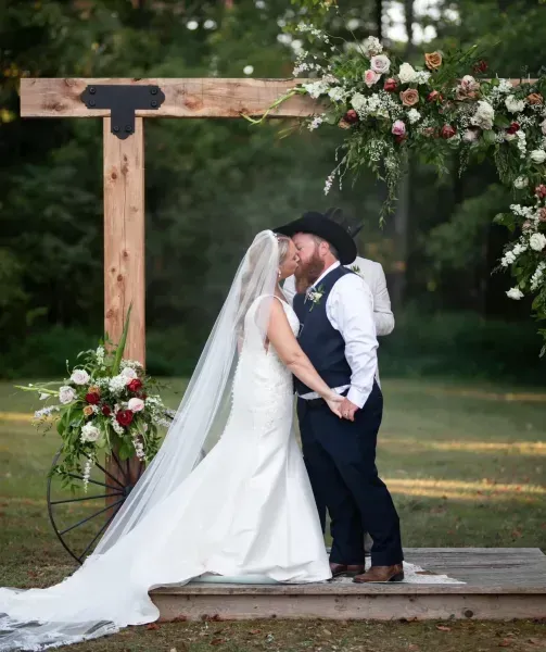 Bride and groom kissing under a wooden floral arch at an outdoor wedding